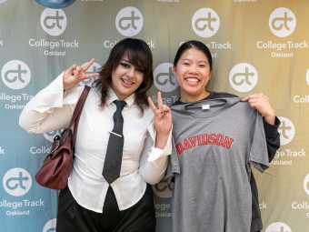 Two smiling students pose in front of a College Track Oakland backdrop, with one student holding up a grey T-shirt featuring "DAVIDSON" in red arched text.
