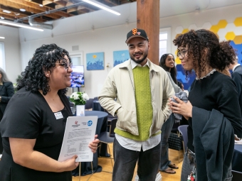 A woman in a black dress holds a Davidson College and College Track partnership document while smiling and speaking with two guests in a bright, modern event space.