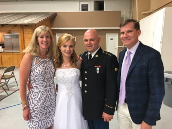 A group of four smiling people pose together indoors, featuring a woman in a patterned sleeveless dress, a woman in a white embellished formal gown, a man in a U.S. Army dress uniform, and a man in a blue plaid blazer with a light purple tie.