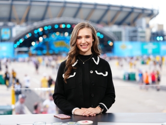 Courtney smiles at a desk with a stadium and large outdoor event stage blurred in the background.