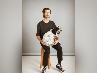 A man with a mustache sits on a wooden stool against a gray backdrop, wearing a black t-shirt, black pants, and black high-top sneakers while cradling a fluffy, black-and-white speckled dog on his lap.