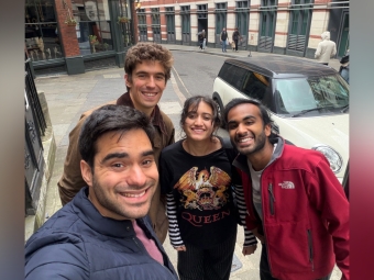Four smiling friends take a high-angle selfie on a curved London street in front of a white compact car and a building labeled "Pub & Eating House."