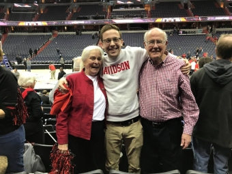 A smiling young man in a white Davidson sweatshirt stands with his arms around an older woman in a red jacket and an older man in a checkered shirt in a basketball arena.