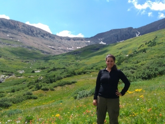 A smiling woman stands in a lush, green mountain valley filled with yellow and red wildflowers under a blue sky with light clouds.