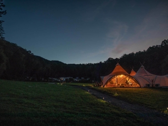 A large, warmly lit event tent sits in a grassy valley at twilight, connected to a gravel path with small ground lights and surrounded by dark, forested hills under a deep blue evening sky.