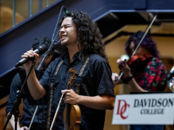 A student with long curly hair sings passionately into a microphone while holding a violin, performing in front of a Davidson College sign.