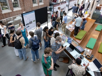 An overhead view of a busy atrium where students and faculty gather around research posters and technical demonstrations during a symposium.