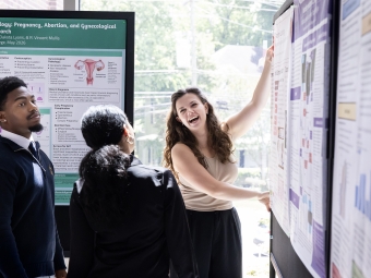 A student laughs while presenting a research poster to two attentive peers.