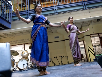 Two students in traditional saris perform a synchronized dance on a stage equipped with drums and microphones.