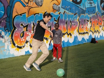 An adult man plays soccer with a young boy in front of a colorful graffiti mural