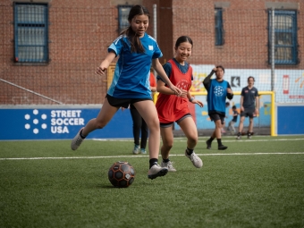 Two young women with dark hair and red and blue team jerseys run with the ball