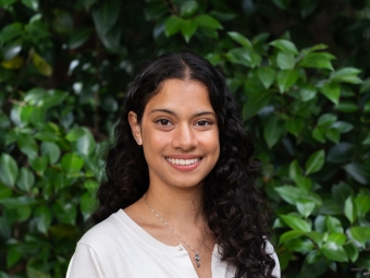 A smiling young woman with long, curly dark hair looks directly at the camera while wearing a white button-up cardigan in front of a dense green leaf background.