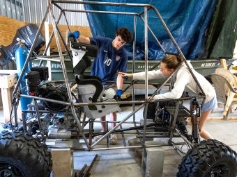 Two students work inside a workshop on a custom Baja-style vehicle frame, with one pointing inside the chassis while the other positions a metallic racing seat.