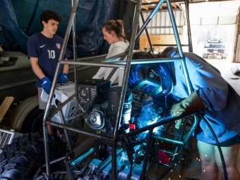 A student wearing a blue welding helmet and protective gloves welds a component onto the metal chassis of a custom off-road vehicle while two peers observe.