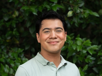 A smiling young man with short dark hair tilts his head slightly while posing in a light green polo shirt against a background of lush green foliage.