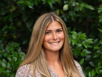 A smiling young woman with long, straight blonde-highlighted hair poses facing forward, wearing a purple and white floral patterned blouse in front of a green leafy backdrop.