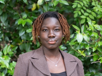 A young woman with reddish-brown twisted locks smiles gently at the camera, wearing a brown blazer over a black top against a background of green bushes.