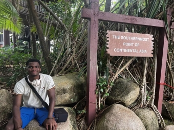 A young man sits on large rocks next to a wooden signpost reading "The Southernmost Point of Continental Asia," surrounded by tropical foliage.