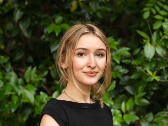 A young woman with blonde hair tied back and loose face-framing strands smiles slightly in a profile pose, wearing a black short-sleeved top against a green leafy backdrop.