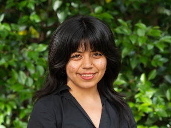 A smiling young woman with dark, shoulder-length layered hair with bangs poses facing forward in a black button-down shirt in front of lush green foliage.