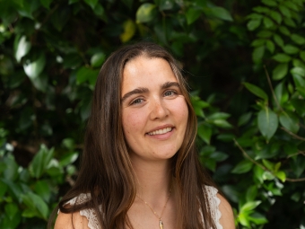 A smiling young woman with long, straight brown hair poses angled slightly toward the camera, wearing a white lace-trimmed tank top against a backdrop of green leaves.