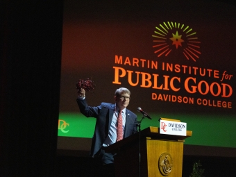 A man in a suit stands at a Davidson College podium on a dimly lit stage, raising a pom-pom in one hand beneath a large projection screen that reads "Martin Institute for Public Good."