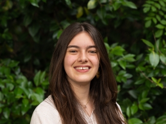 A brightly smiling young woman with long, straight dark brown hair poses facing forward in a cream-colored ribbed long-sleeve shirt against a background of dense green foliage.