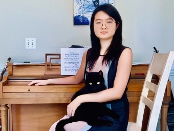A female student with long black hair and glasses sits in front of a piano, holding a black cat