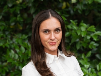A young woman with long, straight dark hair parted down the middle smiles subtly at the camera, wearing a white short-sleeved button-up shirt in front of a green leaf backdrop.