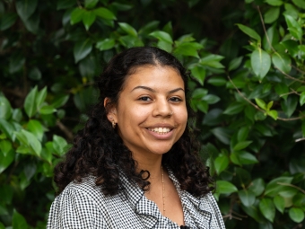 A smiling young woman with long, curly dark hair pinned back at the sides poses at an angle, wearing a black-and-white checkered blouse with a ruffled collar against a leafy green background.