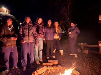 A bundled-up group of students at night around a campfire