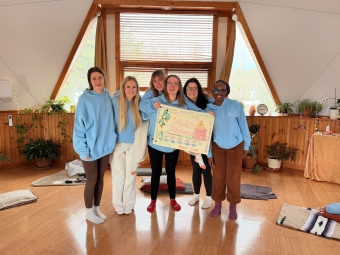 Barefoot students hold a hand-drawn poster in a brightly lit meditation room