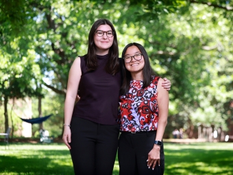 Two smiling students wearing glasses stand side-by-side with arms around each other on a sunny, green campus lawn surrounded by trees.