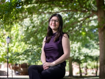 A smiling student wearing glasses and a dark purple sleeveless top sits outside on a stone ledge beneath leafy green trees.