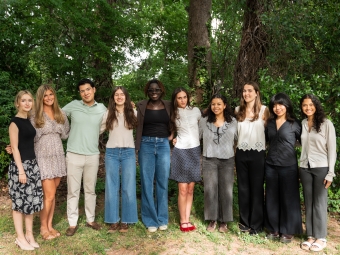 A group of ten young adults stand side by side and smile for a group portrait outdoors in front of a dense backdrop of green trees and foliage.