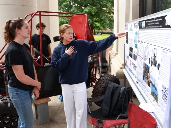 A student points to details on a research presentation board while explaining their engineering project to a peer next to a red Baja vehicle frame.