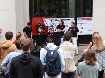 A group of students present their custom red off-road vehicle and research posters to an outdoor audience, with one student speaking into a microphone.