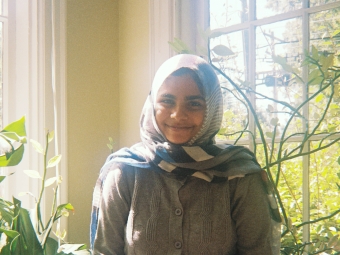 A smiling young woman wearing a patterned hijab and a grey button-up cardigan sits in a sunlit room filled with green plants.