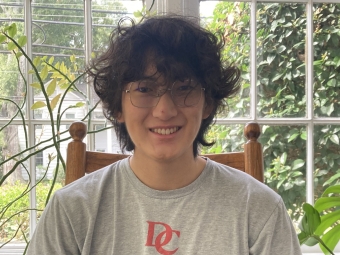 A smiling young man with wavy dark hair and glasses sits in a wooden chair indoors, wearing a grey t-shirt that reads "Davidson College."