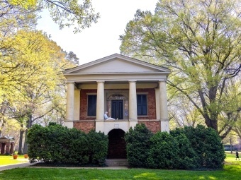 A person sits reading on the upper portico of a historic brick building with classical white columns, surrounded by leafy green trees and hedges on a sunny campus.