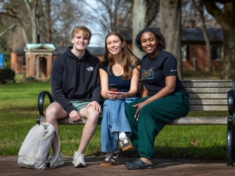 Three students sitting together on a wooden park bench, smiling at the camera.