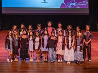 A group of seventeen Davidson College graduates wearing matching black and red ASAPI graduation stoles pose together for a group photo on a theater stage.