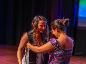 A smiling woman adjust a black graduation stole on a graduate wearing a white dress during a stage celebration.
