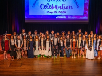 A group of graduates wearing black stoles pose together on a wooden stage in front of a projection screen reading "Latinx Senior Celebration, May 15, 2026."