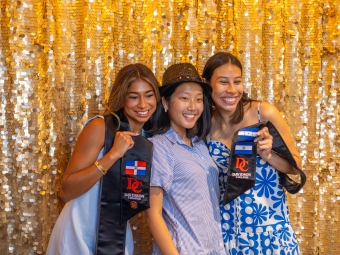 Three smiling young women pose closely together for a photo in front of a glittering gold sequin backdrop, with two displaying their Davidson College graduation stoles.