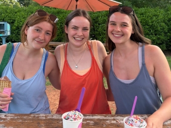 Three young women sitting together at a wooden picnic table under a large orange patio umbrella, holding cups of ice cream topped with colorful sprinkles.