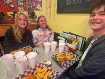 Three young adults smiling at a diner booth table set with burgers, chips, and large drink cups.