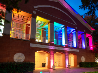 The brick exterior of the Katherine and Tom Belk Visual Arts Center illuminated at dusk with a rainbow gradient of lights across its upper pillars and glowing orbs along the front walkway.