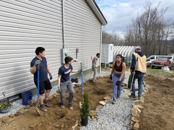 A group of volunteers works together on an outdoor landscaping project next to a house, digging trenches and laying gravel and stone borders.