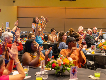 A diverse audience seated at round banquet tables claps, cheers, and raises their hands enthusiastically during an indoor event.